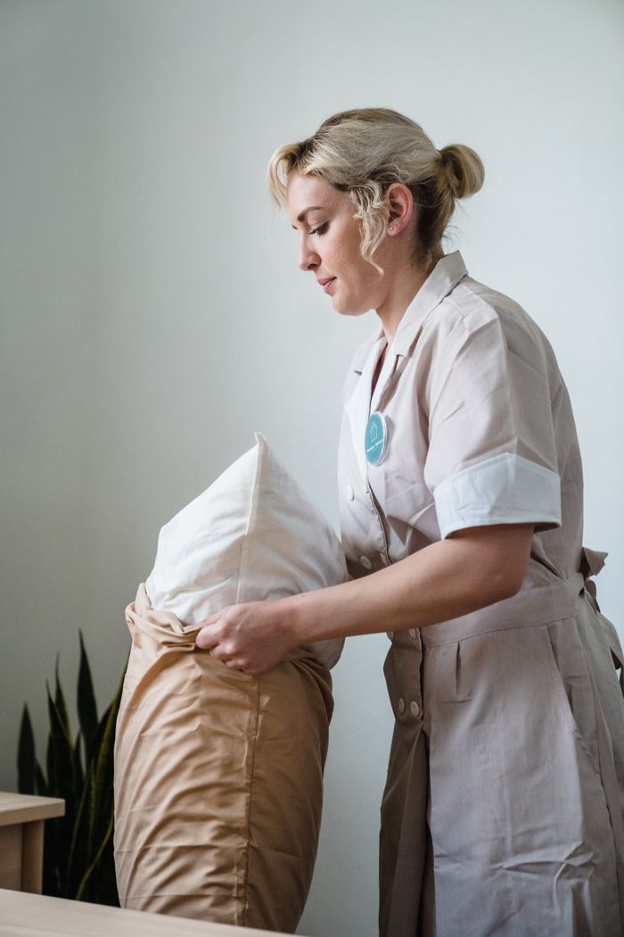 who-we-are Hotel maid in uniform arranging pillows in a neatly styled guest room.